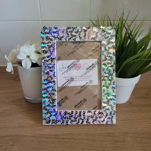 May include: A white picture frame with a black and iridescent sequin border. The frame is decorated with a business card that reads "Jennifer Drew Crafts" and includes a website address.