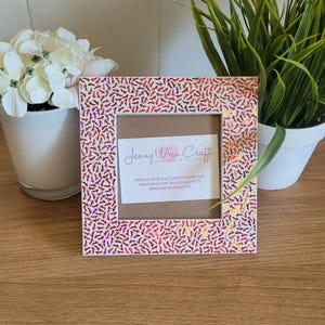 Peut inclure: Un cadre photo blanc avec un motif de paillettes rouge et rose. Le cadre a un passe-partout brun avec le texte "Jenny Wren Craft Handmade with Love" et des informations sur le site web.