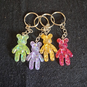 May include: Four bear-shaped keychains made of translucent resin filled with colourful glitter. Each bear features a different hue: lime green, light purple, golden yellow, and bright pink. Each charm is attached to a silver metal split ring.