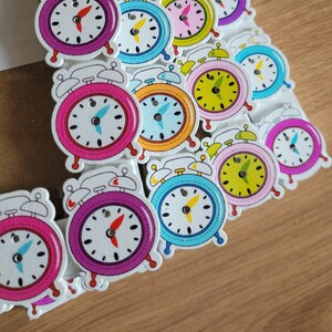 May include: A collection of colorful wooden alarm clock ornaments. The clocks are painted in various colors, including pink, blue, green, yellow, and red. Each clock has a white face with black hands and numbers.