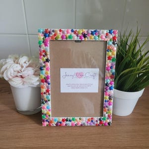 May include: White picture frame with a colorful star design. The frame is decorated with iridescent stars of various colors, including pink, blue, green, and purple. The frame is 4x6 inches and has a white mat.