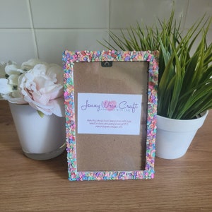 May include: A white picture frame with a colorful sprinkle border. The frame is decorated with pink, blue, green, and yellow sprinkles. The frame is sitting on a wooden surface. The text "Jenny Wren Craft Handmade with Love" is visible on the frame.