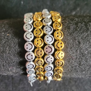 May include: A stack of bracelets with a repeating smiley face design. The bracelets alternate between silver and gold beads, each with a smiling face. The bracelets are stacked on a black surface.