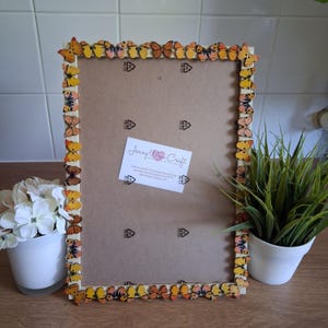 May include: A rectangular picture frame with a light brown backing, decorated with small, colorful butterfly embellishments around the edges. The butterflies are in shades of yellow, orange, and brown. A small white card with text is attached to the frame.