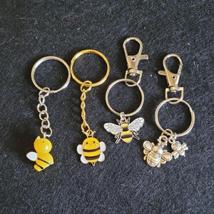 May include: Set of four keychains featuring bee designs. Two keychains have a yellow and black bee charm, one has a yellow bee charm, and one has a silver bee charm with a smaller silver charm.