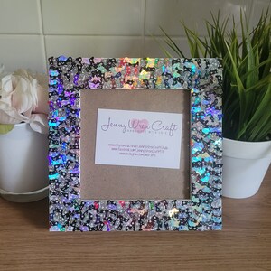 May include: A white picture frame with a black and iridescent glitter border. The frame has a white mat and a card inside with the text "Jenny Wren Craft Handmade with Love" and website information.