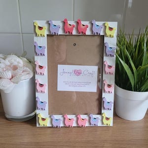 May include: White picture frame with a light brown mat and a white background. The frame is decorated with colorful llama figurines around the border. The frame has a business card inside with the text "Jenny Wren Craft Handmade with Love" and website information.