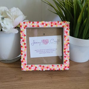 May include: A square picture frame adorned with tiny heart-shaped decorations in red, pink, and yellow. The frame has a white border and a brown backing. The text "Jenny Wren Craft" is visible on a card inside the frame.