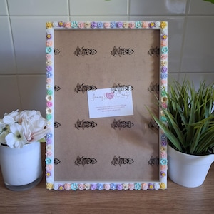 May include: A white picture frame decorated with small, colorful flowers. The frame has a rectangular shape and a neutral background. A small card with text is attached to the frame. A small potted plant and flowers are on either side of the frame.