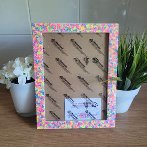 May include: A white picture frame with a pink, blue and yellow glitter border. The frame is sitting on a wooden surface. The frame is empty, but there is a small business card inside.