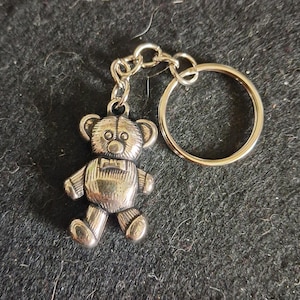 May include: A silver-toned metal keyring featuring a small, textured teddy bear charm attached to a circular split ring by a short chain. The bear has detailed etched lines on its body, rounded ears, and a small bow tie detail on its chest.