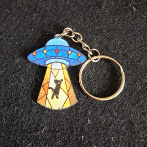May include: A keychain featuring a blue UFO with orange circular accents and a yellow light beam. Inside the beam, a black cat silhouette is depicted. The design has a stained glass appearance with dark outlines and a silver metal ring attached.