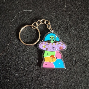 May include: A colorful keychain featuring a cartoon alien in a UFO. The UFO is decorated with flowers and a mushroom, with a rainbow-colored beam. The keychain has a gold-colored ring and chain.