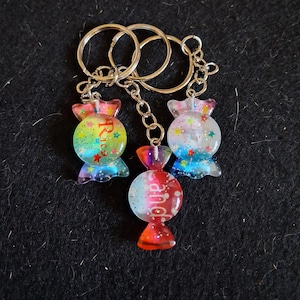 May include: Three candy-shaped keychains with silver keyrings. Each keychain features a colorful, glitter-filled candy design with the word "Rice" or "and" on the front. The candies have a translucent appearance with red, blue, and purple accents.