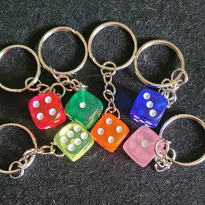 May include: A selection of dice keyrings in various colours. The dice are red, green, orange, yellow, blue, and pink, each with white dots. The keyrings have silver-coloured metal rings and chains.