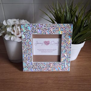 May include: Square picture frame with a white border decorated with iridescent dog bone shapes. The frame has a brown backing and a white label with the text "Jenny Wren Craft". The frame is sitting on a wooden surface.