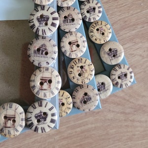 May include: A pale blue picture frame adorned with vintage buttons. The buttons display clock faces, crowns, and architectural landmarks. The frame is on a light brown surface.