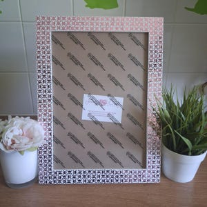 May include: A rectangular picture frame with a metallic rose gold finish featuring a repeating pattern of small skulls. The frame contains a brown protective sheet with repeating text and a central white label with printed text and a logo.