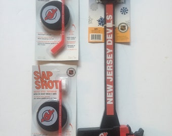 May include: Three New Jersey Devils hockey themed items: a red and black toothbrush with a hockey stick handle, a black and red ice scraper with a Zamboni design, and a black and red hockey puck with the Devils logo.