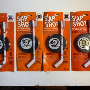 May include: Four hockey-themed toothbrushes with puck-shaped holders. Each package features a different team logo: San Jose Sharks, Mighty Ducks, Buffalo Sabres, and Boston Bruins. The toothbrushes are white with hockey stick handles.