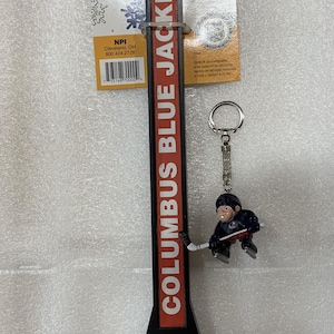 Puede incluir: Un abrebotellas con el tema de los Columbus Blue Jackets con un llavero de un jugador de hockey. El abridor tiene un dise&ntilde;o rojo y negro con el logotipo del equipo y las palabras "COLUMBUS BLUE JACKETS". El llavero presenta un jugador de hockey.