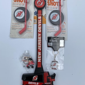 May include: A set of New Jersey Devils hockey memorabilia, including a red and black Zamboni toy, a hockey stick toothbrush, a key chain with a jersey charm, and a puck with the team logo. The items are packaged in clear plastic bags and are ready for a fan to enjoy.