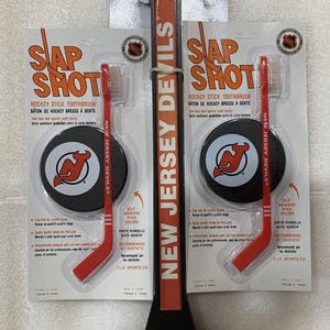Puede incluir: Un soporte para cepillo de dientes de hockey negro y rojo con un disco autoadhesivo. El soporte presenta el logotipo de los New Jersey Devils y las palabras "New Jersey Devils" y "Slap Shot".