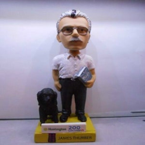 May include: A bobblehead figurine of James Thurber, a white male with glasses and a mustache, wearing a white shirt and black pants, holding a book. The figurine is standing on a base with the text "Huntington 200 Columbus" and "JAMES THURBER". A black dog figurine is standing to the left of the bobblehead.