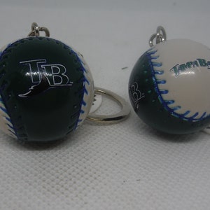 Könnte beinhalten: Zwei grün-weiße Baseball-Schlüsselanhänger mit dem Logo der Tampa Bay Rays. Ein Schlüsselanhänger zeigt das Logo des Teams, der andere die Worte "Tampa Bay".