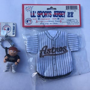 May include: A white and blue baseball jersey with the word "Astros" in brown lettering. The jersey is a keychain with a small plastic baseball player figure attached.