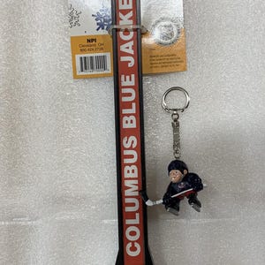 Puede incluir: Un abrebotellas con el tema Zamboni de los Columbus Blue Jackets con un llavero de jugador de hockey. El abridor es rojo y negro con "COLUMBUS BLUE JACKETS" en blanco. El llavero presenta una figura de jugador de hockey.