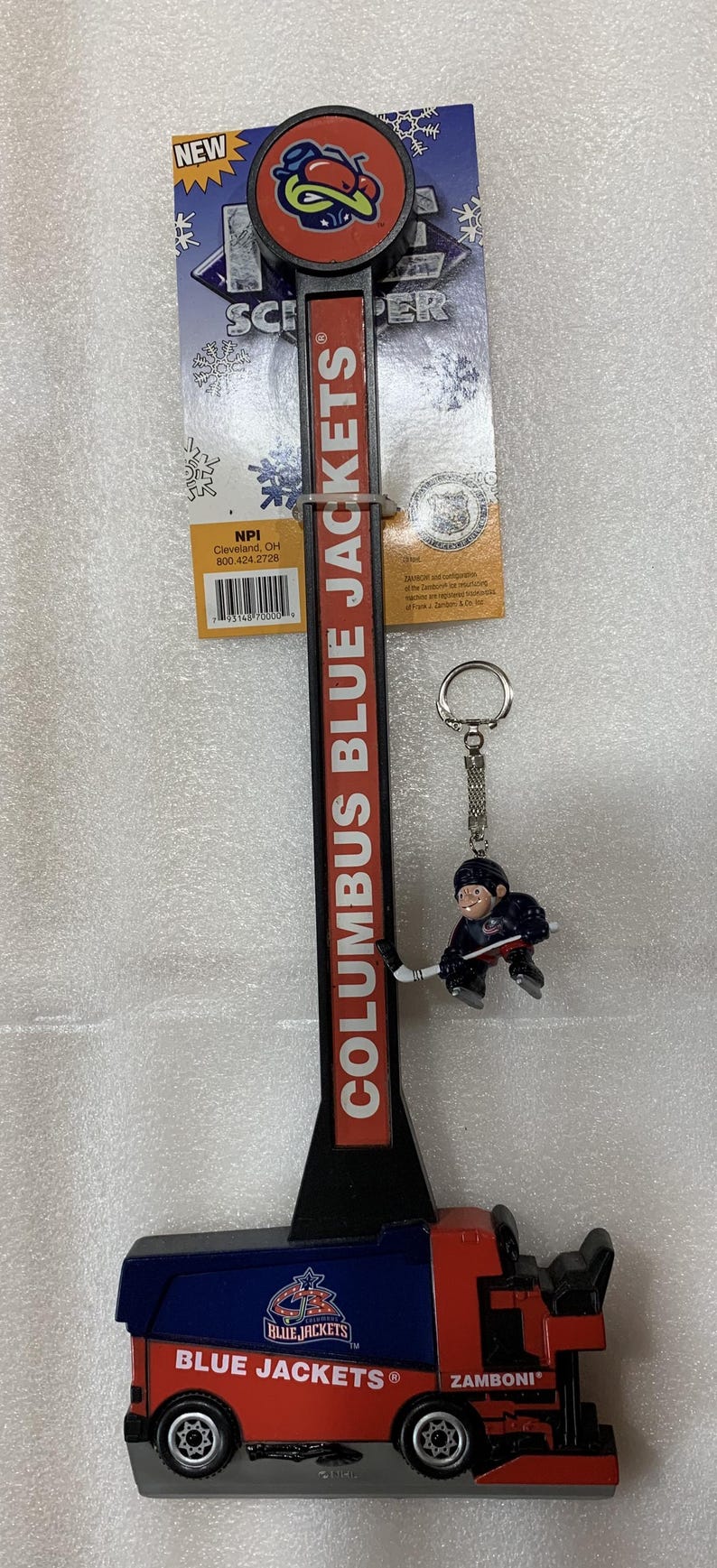 Puede incluir: Un abrebotellas con el tema Zamboni de los Columbus Blue Jackets con un llavero de jugador de hockey. El abridor es rojo y negro con "COLUMBUS BLUE JACKETS" en blanco. El llavero presenta una figura de jugador de hockey.