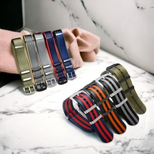 May include: Assortment of colorful watch straps in various colors, including green, gray, blue, red, orange, and black. The straps are made of nylon and have a woven design.