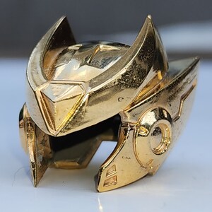 K&ouml;nnte beinhalten: Ein goldfarbener Helm mit einem detaillierten Design. Der Helm hat eine spitze Oberseite und ein gebogenes Visier. Der Helm ist aus Metall und hat eine gl&auml;nzende Oberfl&auml;che.
