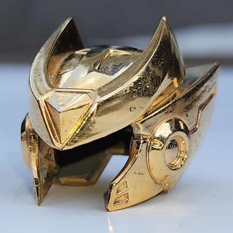 K&ouml;nnte beinhalten: Ein goldfarbener Helm mit einem detaillierten Design. Der Helm hat eine spitze Oberseite und ein gebogenes Visier. Der Helm ist aus Metall und hat eine gl&auml;nzende Oberfl&auml;che.