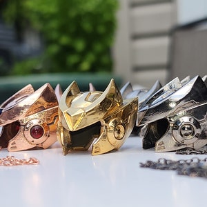 K&ouml;nnte beinhalten: Drei Metallanh&auml;nger in Form eines Roboterkopfes. Die Anh&auml;nger sind in Gold, Silber und Ros&eacute;gold gehalten. Jeder Anh&auml;nger hat eine Kette daran.