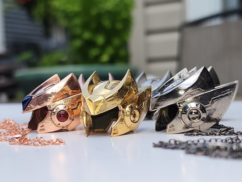 K&ouml;nnte beinhalten: Drei Metallanh&auml;nger in Form eines Roboterkopfes. Die Anh&auml;nger sind in Gold, Silber und Ros&eacute;gold gehalten. Jeder Anh&auml;nger hat eine Kette daran.