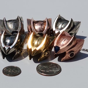 K&ouml;nnte beinhalten: F&uuml;nf Metallanh&auml;nger in Helmform, jeder mit einer anderen Oberfl&auml;che: Silber, Gold, Kupfer, Bronze und Ros&eacute;gold. Die Anh&auml;nger sind in einem Halbkreis angeordnet, mit einer M&uuml;nze davor.