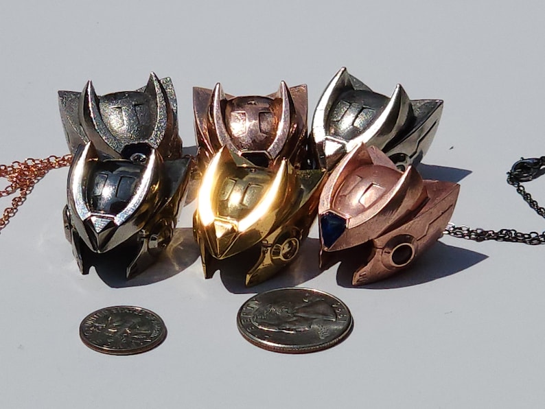 K&ouml;nnte beinhalten: F&uuml;nf Metallanh&auml;nger in Helmform, jeder mit einer anderen Oberfl&auml;che: Silber, Gold, Kupfer, Bronze und Ros&eacute;gold. Die Anh&auml;nger sind in einem Halbkreis angeordnet, mit einer M&uuml;nze davor.