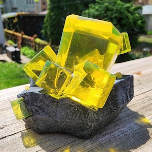 Könnte beinhalten: Eine dekorative Skulptur mit leuchtend gelben, geometrischen Kristallformationen, die aus einer dunkelgrauen, strukturierten Basis herausragen. Die Kristalle sind durchscheinend und fangen das Licht ein, die Basis hat eine raue, steinartige Oberfläche. Die Skulptur steht auf einer Holzoberfläche.