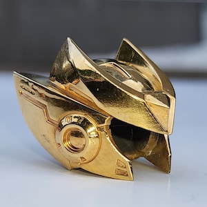K&ouml;nnte beinhalten: Ein goldfarbener Roboterhelm mit einem detaillierten Design. Der Helm hat ein gro&szlig;es, rundes Visier und einen spitzen Kamm.