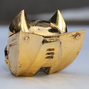 K&ouml;nnte beinhalten: Ein goldfarbener Metallhelm mit einer spitzen Oberseite und zwei ohrenartigen Vorspr&uuml;ngen. Der Helm hat eine glatte, gl&auml;nzende Oberfl&auml;che und ein detailliertes Design.
