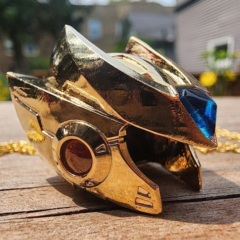 K&ouml;nnte beinhalten: Ein goldfarbener Metallanh&auml;nger in Form eines Helms mit einem blauen Edelstein an der Seite. Der Anh&auml;nger hat eine Kette.