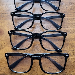 May include: Four pairs of black-framed glasses arranged in a vertical column. Each pair features a classic rectangular shape with clear lenses and small silver-toned metallic accents on the outer corners of the frames near the hinges.