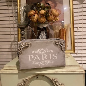 May include: A gray wooden box with the word "PARIS" written in white on the front. The box is decorated with ornate silver accents and sits on a light green table.