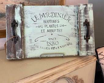 Könnte beinhalten: Ein rustikales Holztablett mit hellgrünem Hintergrund und abgenutzter weißer Farbe. Das Tablett zeigt ein schwarzes Schablonenmuster mit der Aufschrift "Le Jardinier Boutures - Plantes - Et Arbustes Seit 1830 Par Amour De La Nature". Das Tablett hat zwei Metallgriffe an den Seiten.