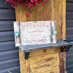 May include: A rustic wooden sign with white paint and red lettering that reads "PATISSERIE Pays de Forcalquier Provence Le Petit Gateau". The sign has two turquoise blue knobs on the top and is sitting on a shelf attached to a weathered yellow door.