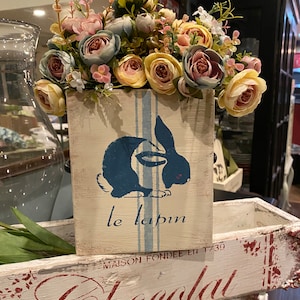May include: A white wooden box with blue stripes and a painted image of a rabbit with the text "le lapin" and "Maison Fondée en 1/39". The box is filled with a bouquet of pastel pink, yellow, and blue flowers. The box is sitting on top of a larger white wooden box with the text "Chocolat" in red.