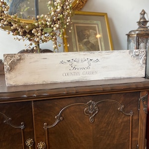 May include: White wooden sign with black lettering that reads "French Country Garden". The sign is distressed and has a rustic look. It is sitting on a dark brown wooden cabinet.