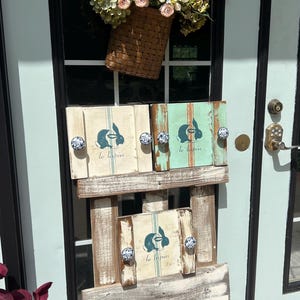 May include: Three wooden signs with a bunny rabbit design and the text "le lapin" on each sign. The signs are displayed on a wooden pallet stand. The signs are painted in shades of blue, green, and white. A basket of pink and white flowers hangs above the signs.